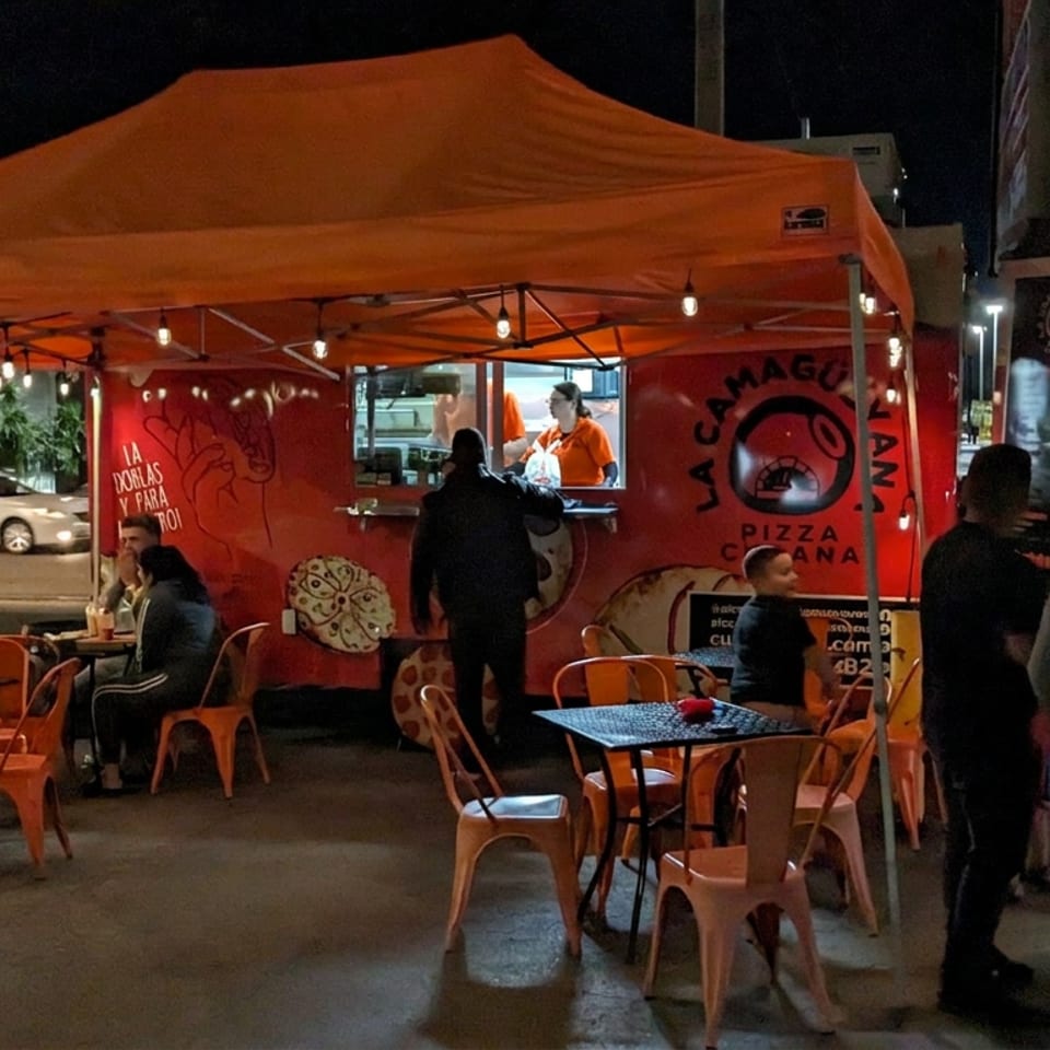 Camión de Pizza Cubana La Camagüeyana con asientos al aire libre y clientes bajo un toldo naranja por la noche.