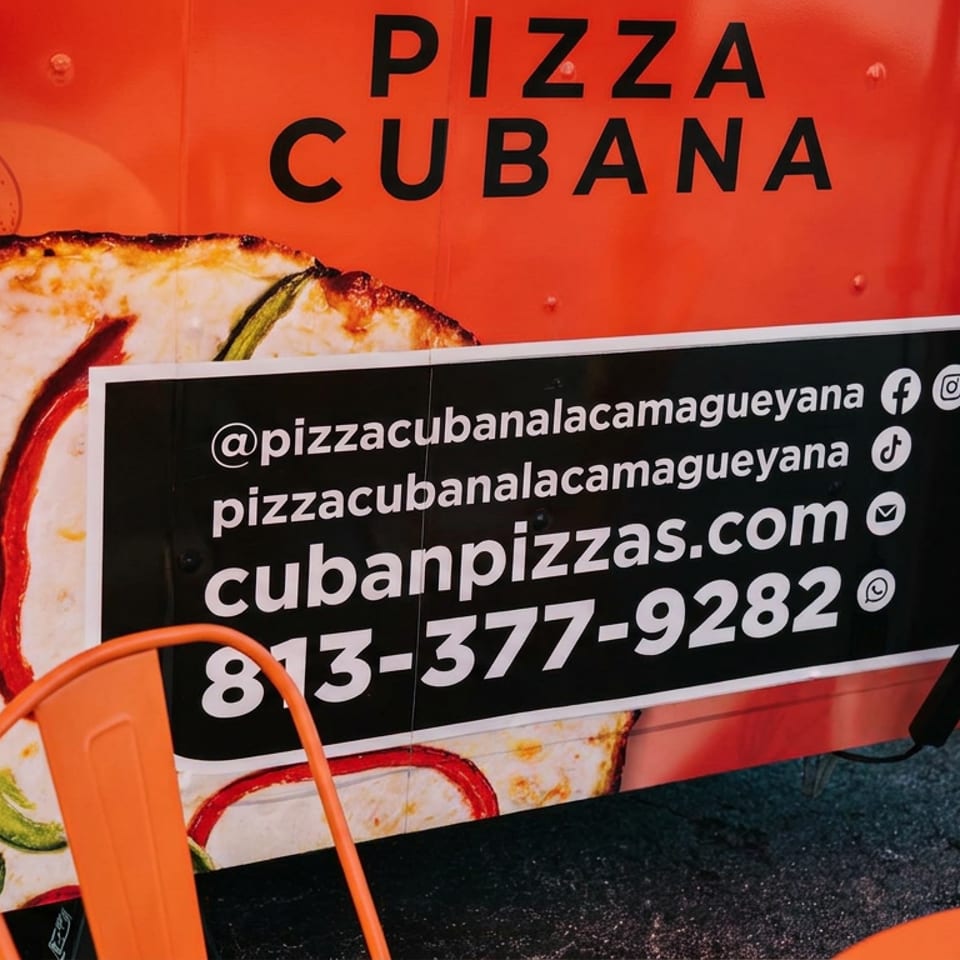 Camión rojo de Pizza Cubana con imagen de pizza y letrero de contacto; hay sillas naranjas cerca.