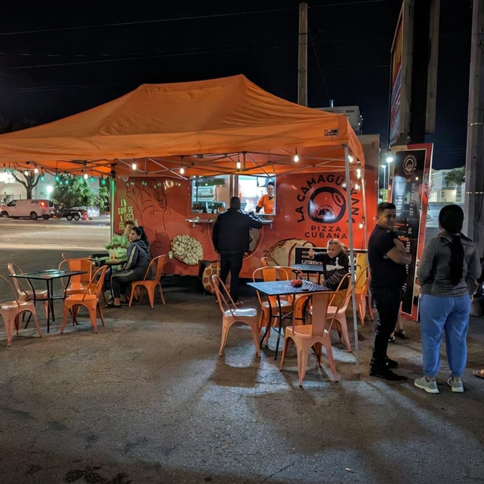 Camión de pizza cubana La Camagüeyana con mesas al aire libre y clientes bajo un toldo naranja por la noche.